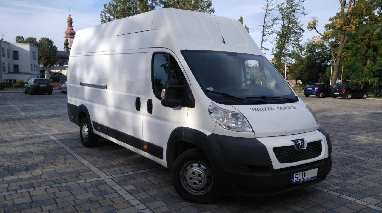 Biały samochód dostawczy Peugeot na parkingu z kostki brukowej, w tle budynki i drzewa.