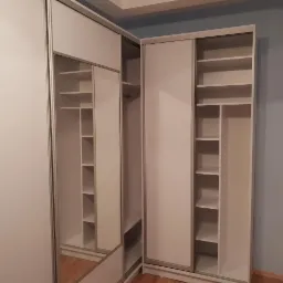 Biała szafa narożna z przesuwnymi drzwiami i lustrem, z widocznymi półkami wewnętrznymi, na tle ściany w odcieniach szarości i beżu.