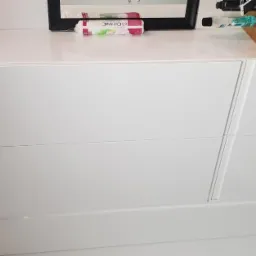 Biała szafka na wymiar z trzema szufladami, widoczne akcesoria na blacie i fragment ściany z cegły.