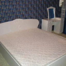 Białe łóżko z dekoracyjnym zagłówkiem i toaletka z lustrem na tle niebieskiej tapety w pokoju.