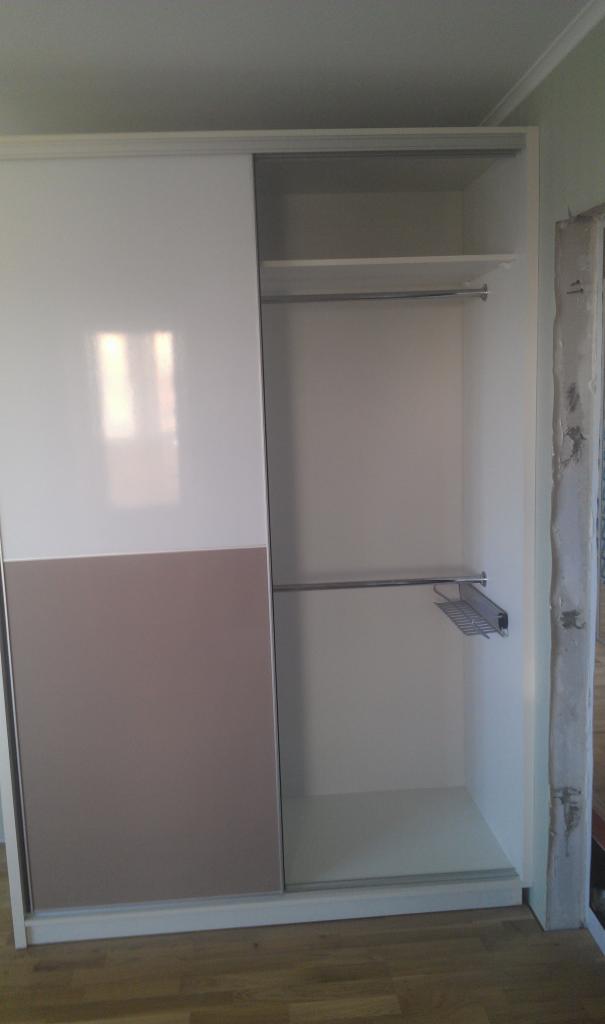 Biała szafa przesuwna z beżowym panelem dolnym, częściowo otwarta, ukazująca puste półki i drążek na wieszaki, widoczny fragment surowego muru w tle.