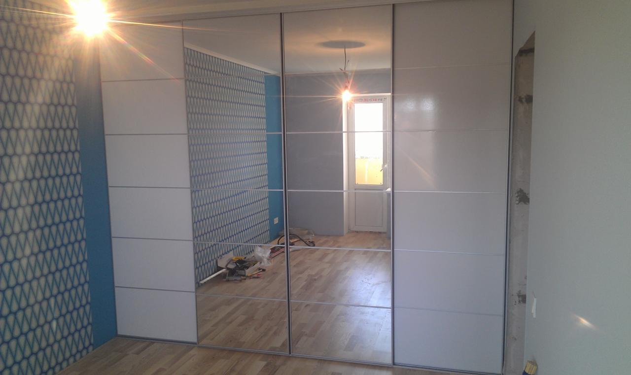 Nowoczesna szafa wnękowa z przesuwnymi drzwiami z lustrami, odbijająca wnętrze pokoju z niebieską tapetą i drewnianą podłogą, widoczne narzędzia i przewody na podłodze sugerują trwający montaż.