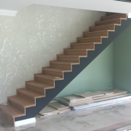 Tomasz Kolodziejczyk - Drewniane schody na czarnej konstrukcji, częściowo zamontowane, z widocznymi paczkami paneli pod schodami i ułożoną podłogą w jodełkę.