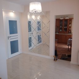 Tomasz Kolodziejczyk - Jasne wnętrze z błyszczącą podłogą, dekoracyjnym lustrem z fazowanych płytek i widokiem na kuchnię z drewnianymi meblami. W rogu widoczne narzędzia.