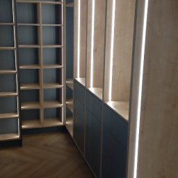 Tomasz Kolodziejczyk - Wnętrze z zabudową meblową: regał z półkami podświetlonymi LED, dolne szafki z ciemnymi frontami, parkiet ułożony w jodełkę.