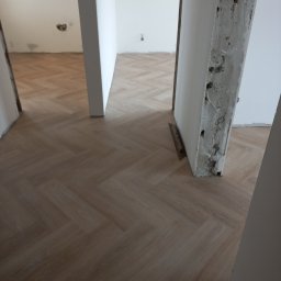 Tomek - Ułożona podłoga z paneli w jodełkę, widoczne surowe obramowania drzwi w trakcie remontu, ślady po instalacjach na ścianach w tle.