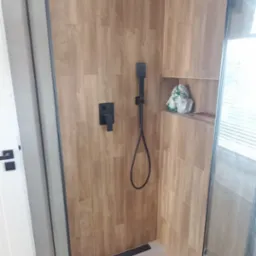 Nowoczesna kabina prysznicowa z deszczownicą i panelami imitującymi drewno. Czarna armatura, minimalistyczny design, jasna podłoga. Wykończenie łazienki w neutralnych barwach.