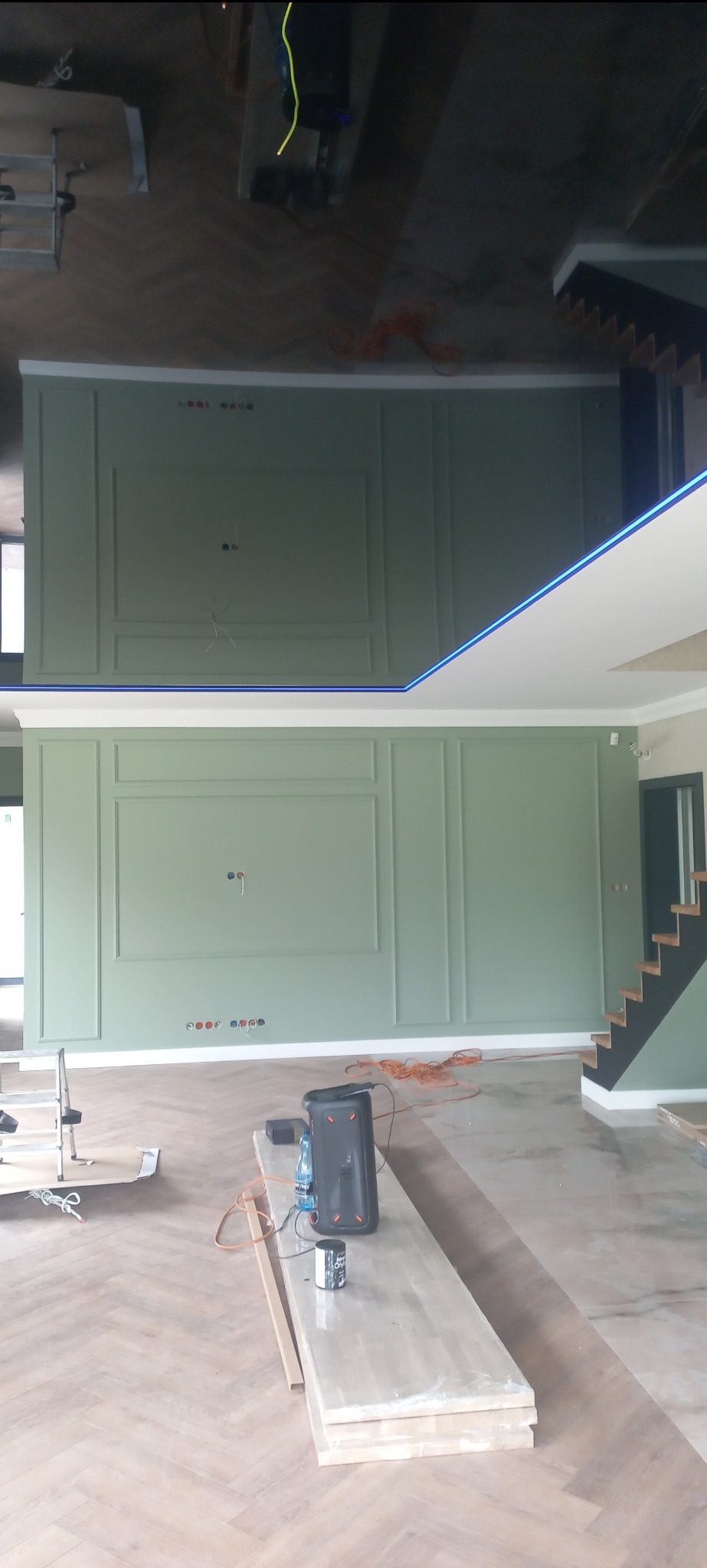 Wnętrze w trakcie prac wykończeniowych: ściany w kolorze szałwiowym z dekoracyjnymi panelami, podłoga ułożona w jodełkę, widoczne kable, drabina, głośnik JBL i niezabezpieczone otwory elektryczne.