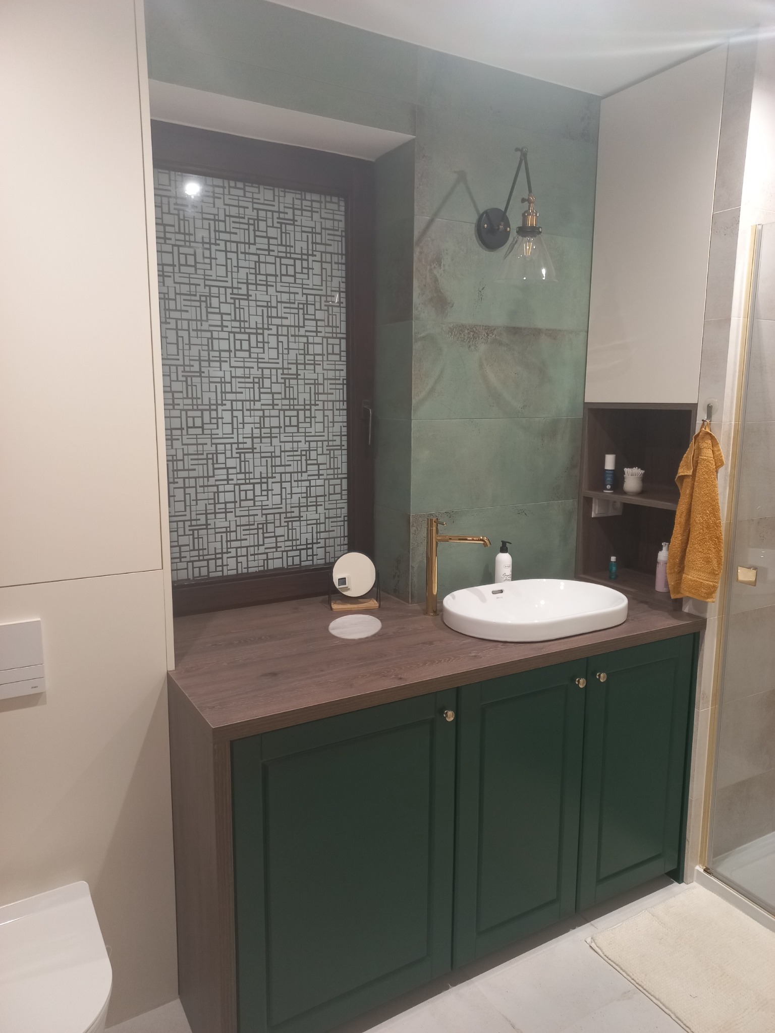 Nowoczesna łazienka w Pabianicach z zielonymi szafkami pod umywalką, drewnianym blatem, złotą baterią i dekoracyjną szybą w oknie.