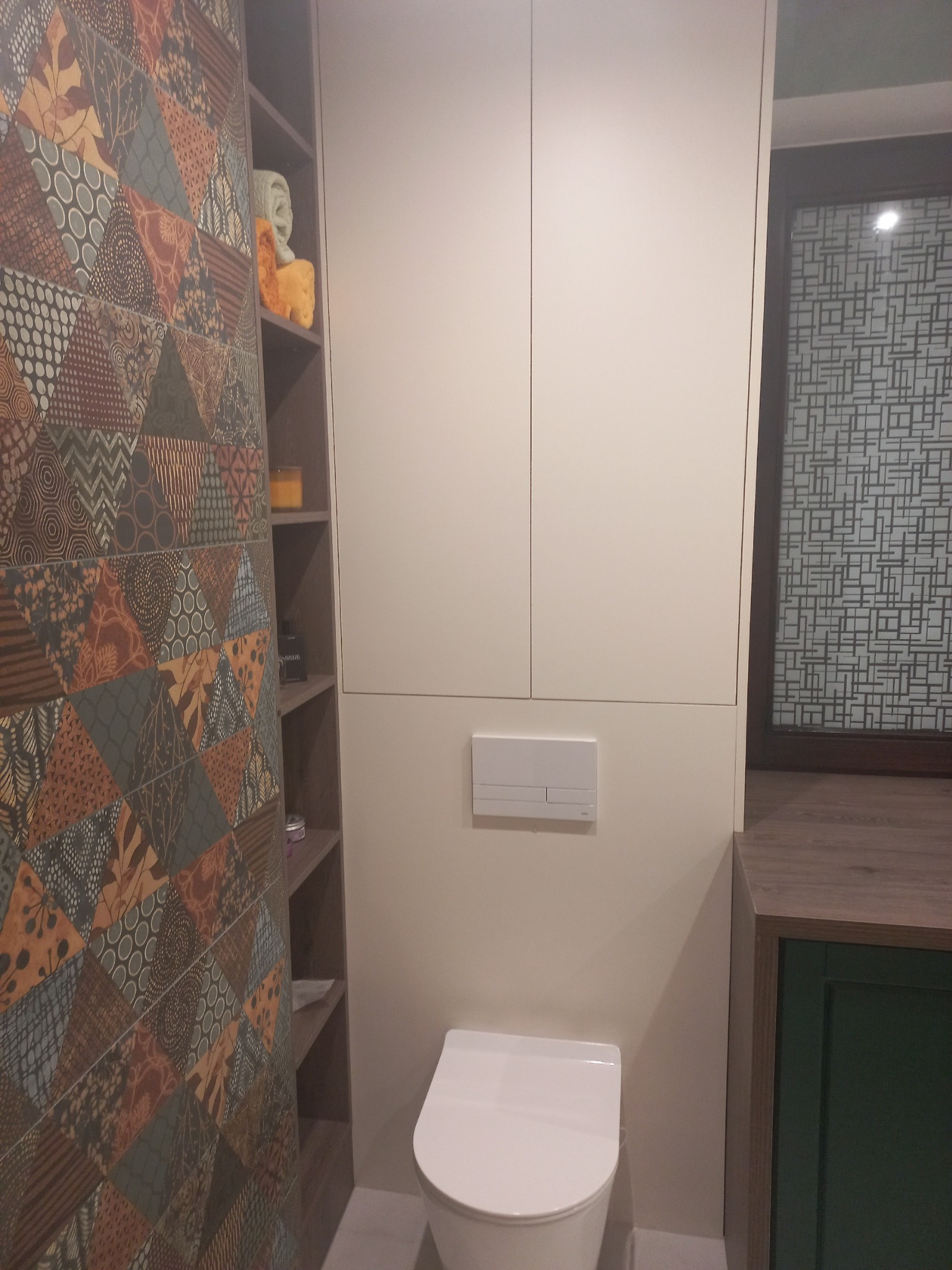 Nowoczesna łazienka z zabudowaną toaletą, szafką nad nią i półkami z dekoracyjną, wzorzystą okładziną ścienną po lewej stronie oraz oknem z geometryczną folią.