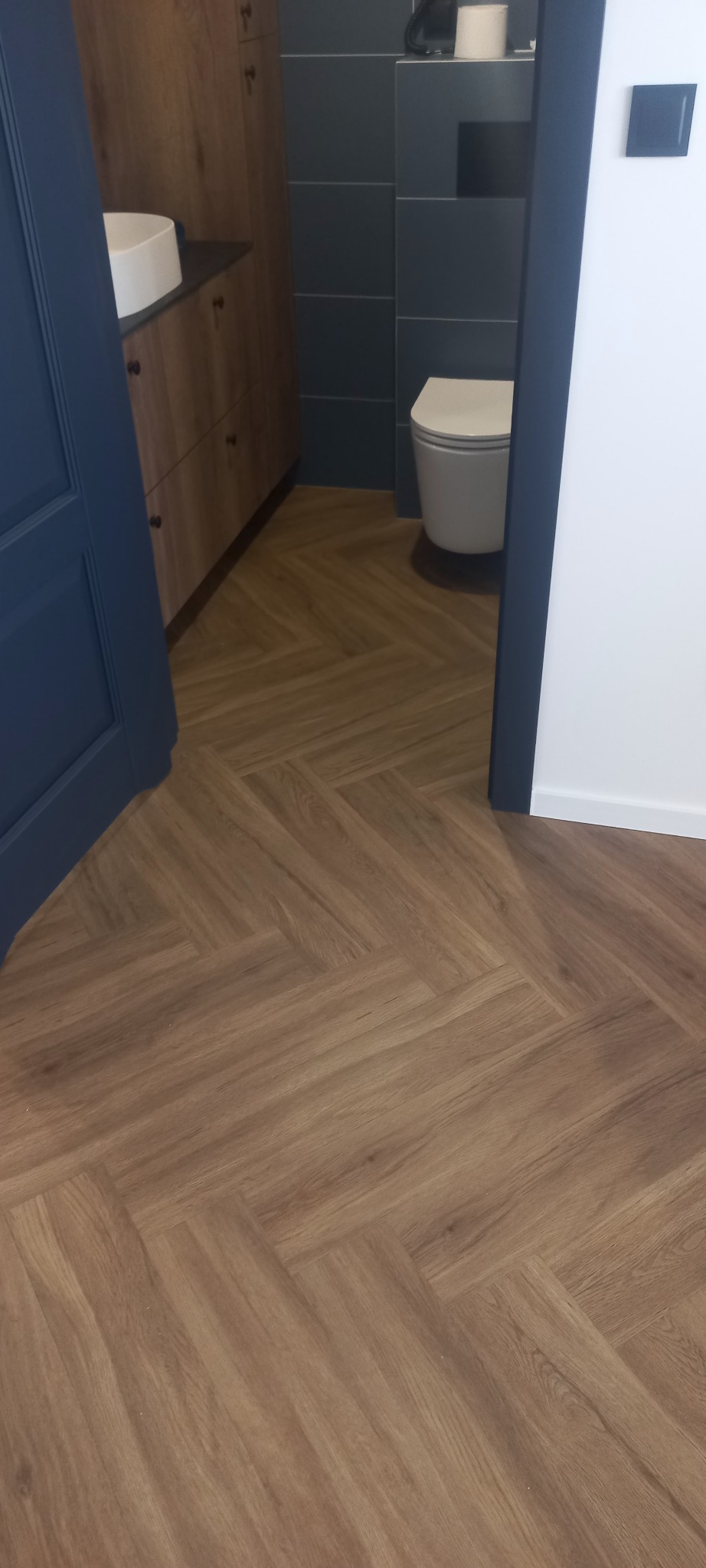 Ułożona podłoga z paneli w jodełkę w łazience z elementami wyposażenia, widok z perspektywy wejścia.