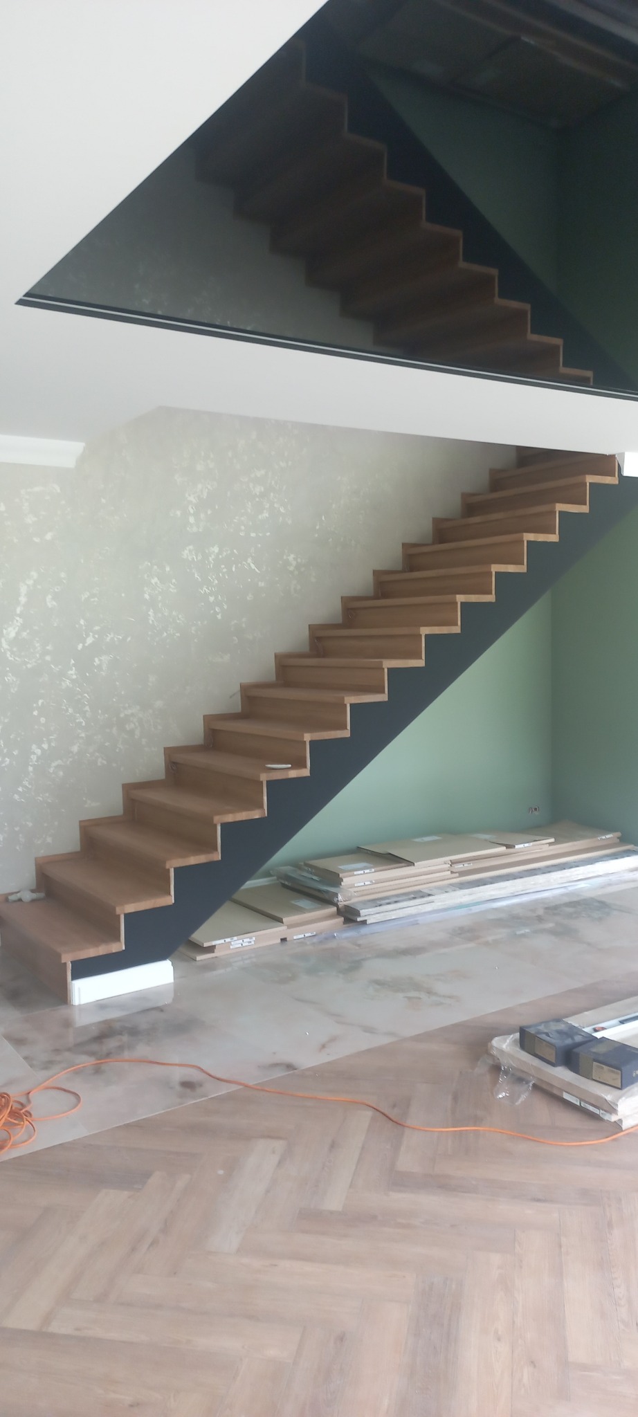 Drewniane schody na czarnej konstrukcji, częściowo zamontowane, z widocznymi paczkami paneli pod schodami i ułożoną podłogą w jodełkę.