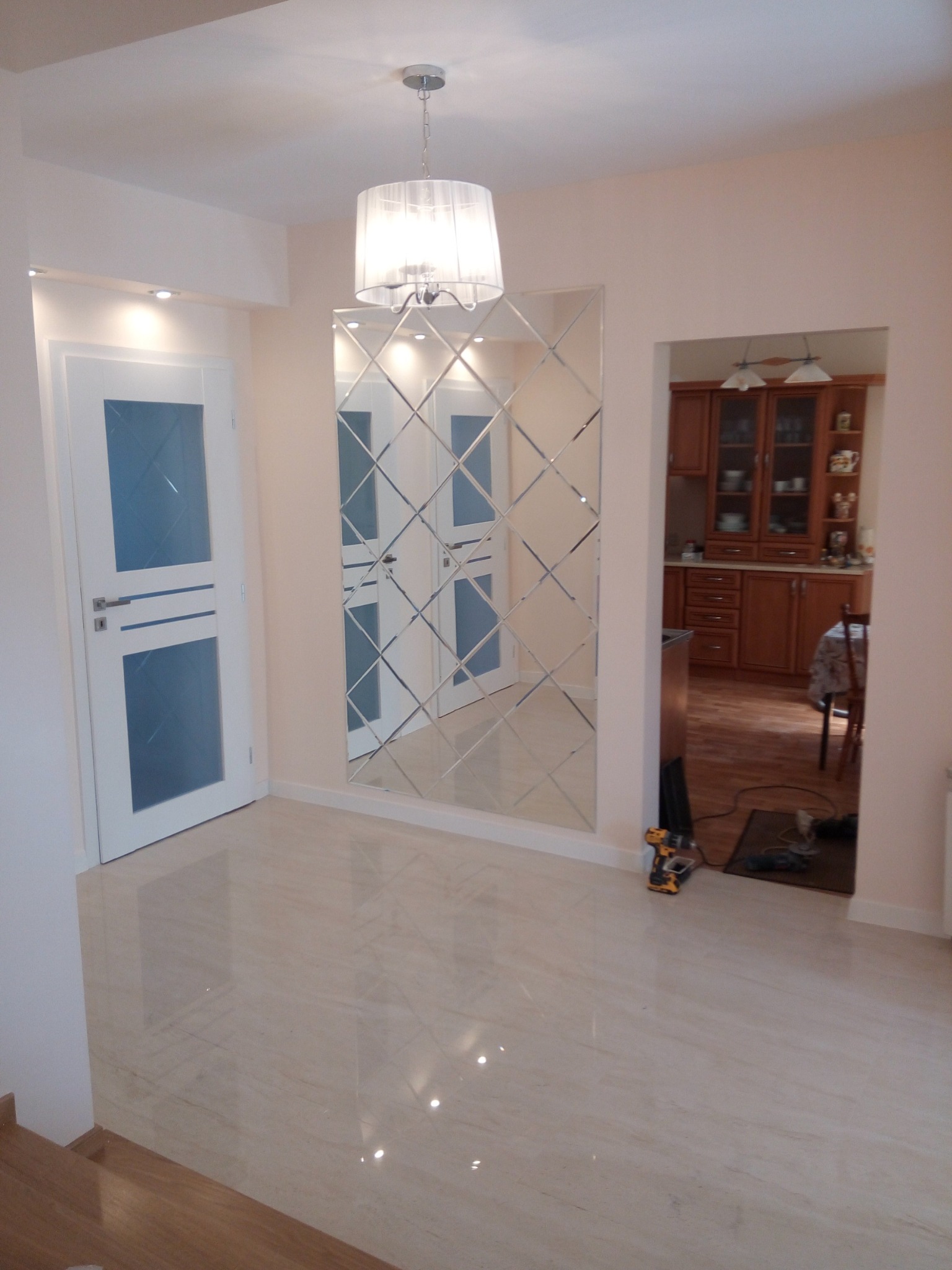 Jasne wnętrze z błyszczącą podłogą, dekoracyjnym lustrem z fazowanych płytek i widokiem na kuchnię z drewnianymi meblami. W rogu widoczne narzędzia.