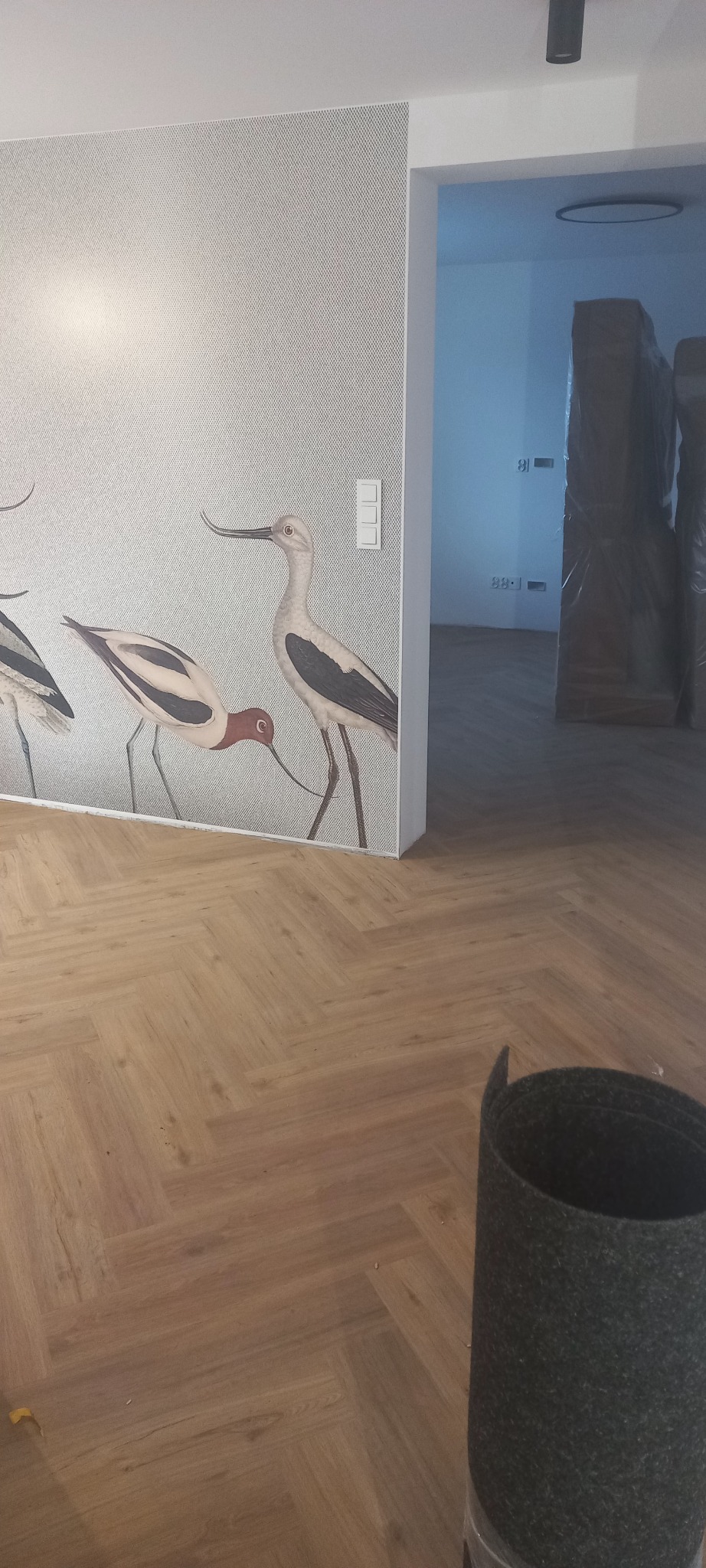 Ułożona podłoga z paneli w jodełkę, na ścianie tapeta z motywem ptaków, w tle widoczne meble zabezpieczone folią