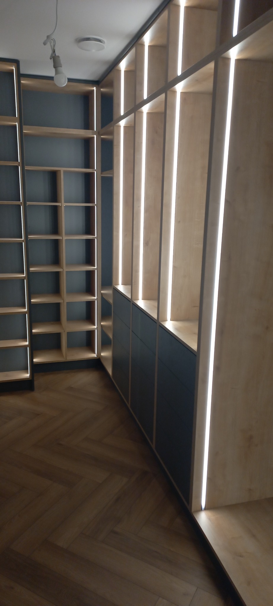 Wnętrze z zabudową meblową: regał z półkami podświetlonymi LED, dolne szafki z ciemnymi frontami, parkiet ułożony w jodełkę.