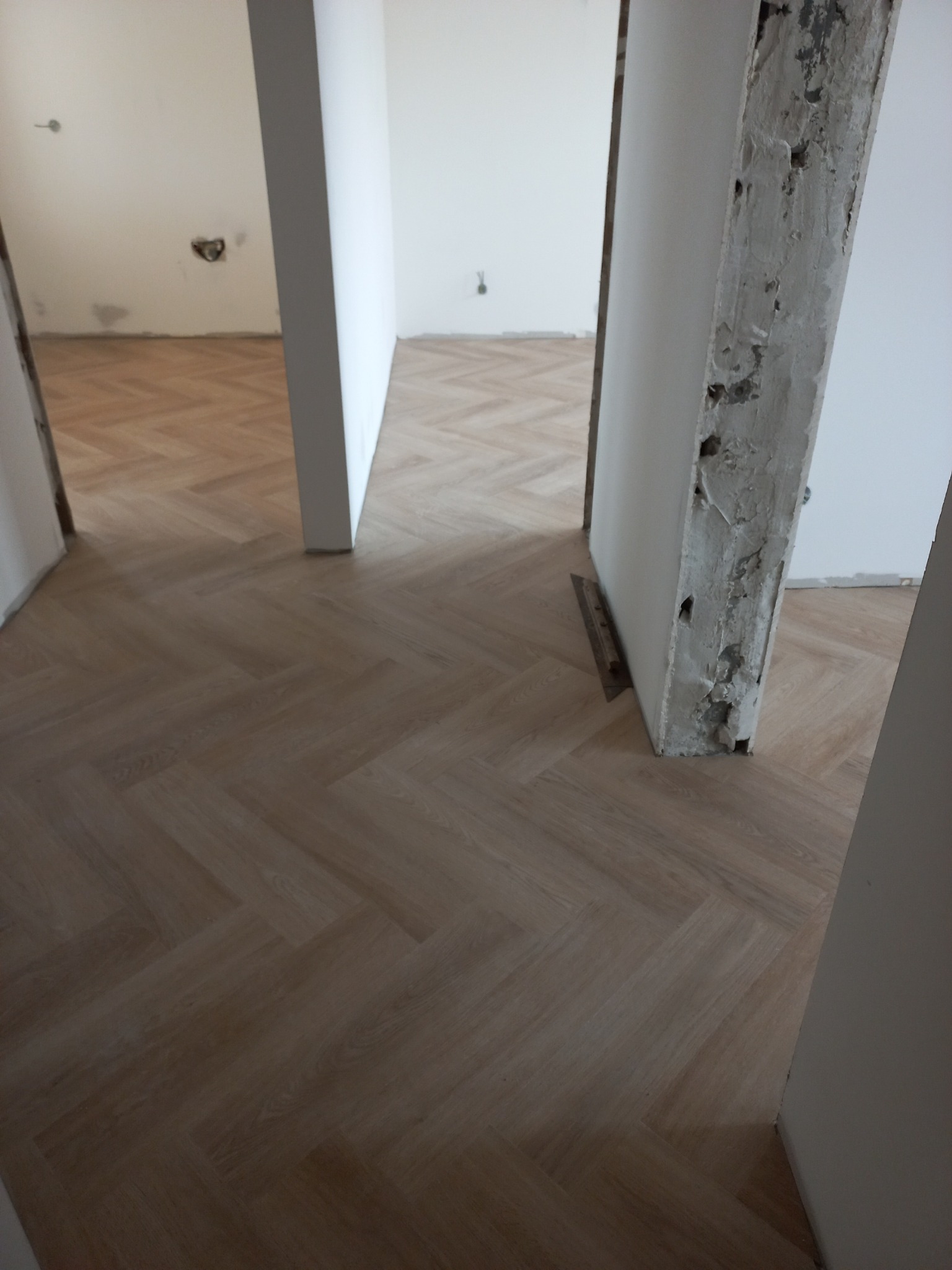 Ułożona podłoga z paneli w jodełkę, widoczne surowe obramowania drzwi w trakcie remontu, ślady po instalacjach na ścianach w tle.