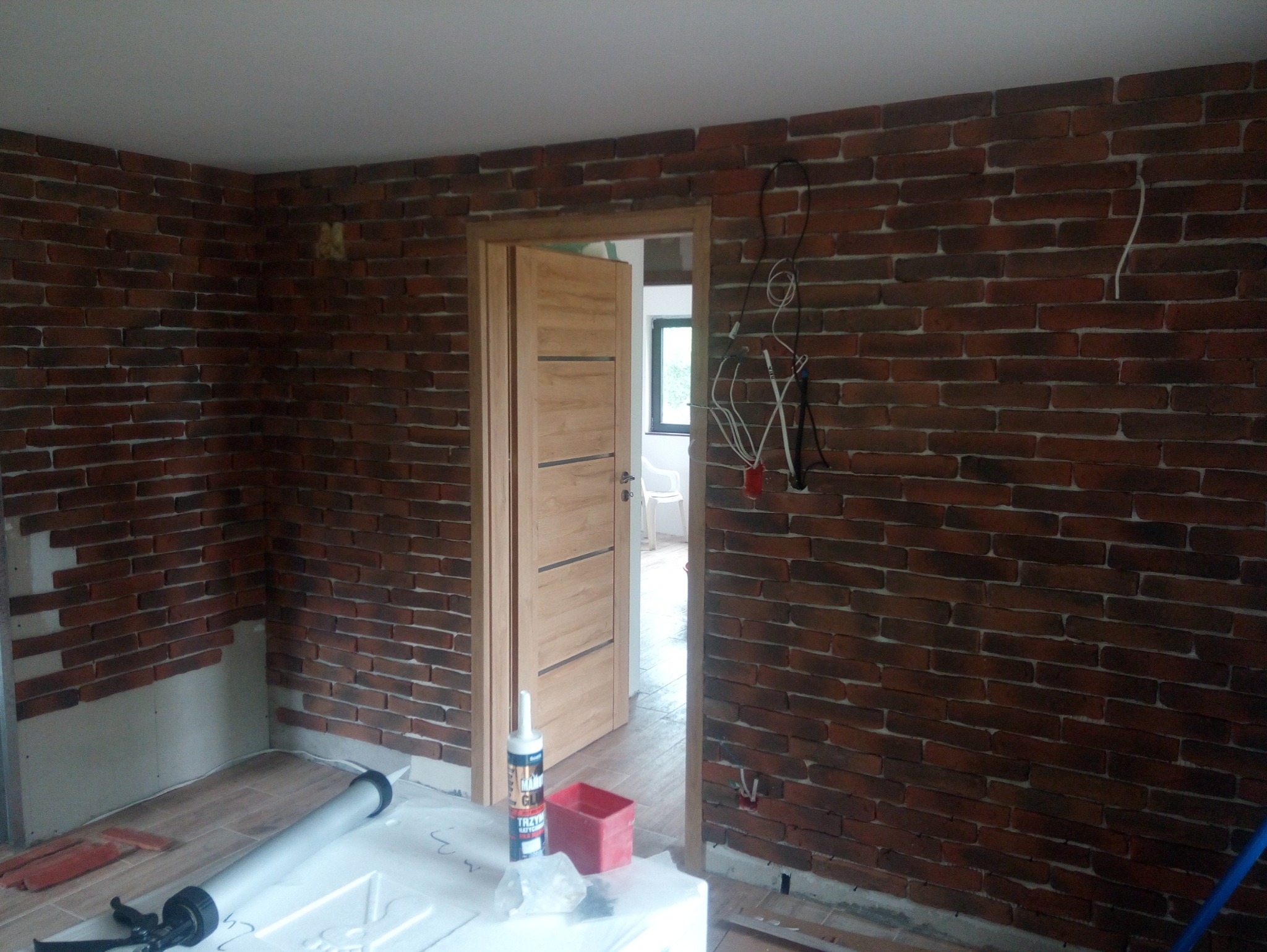 Pokój w trakcie wykańczania z imitacją ceglanej ściany, widoczne narzędzia i materiały budowlane, częściowo ułożona podłoga, otwarte drewniane drzwi do kolejnego pomieszczenia.