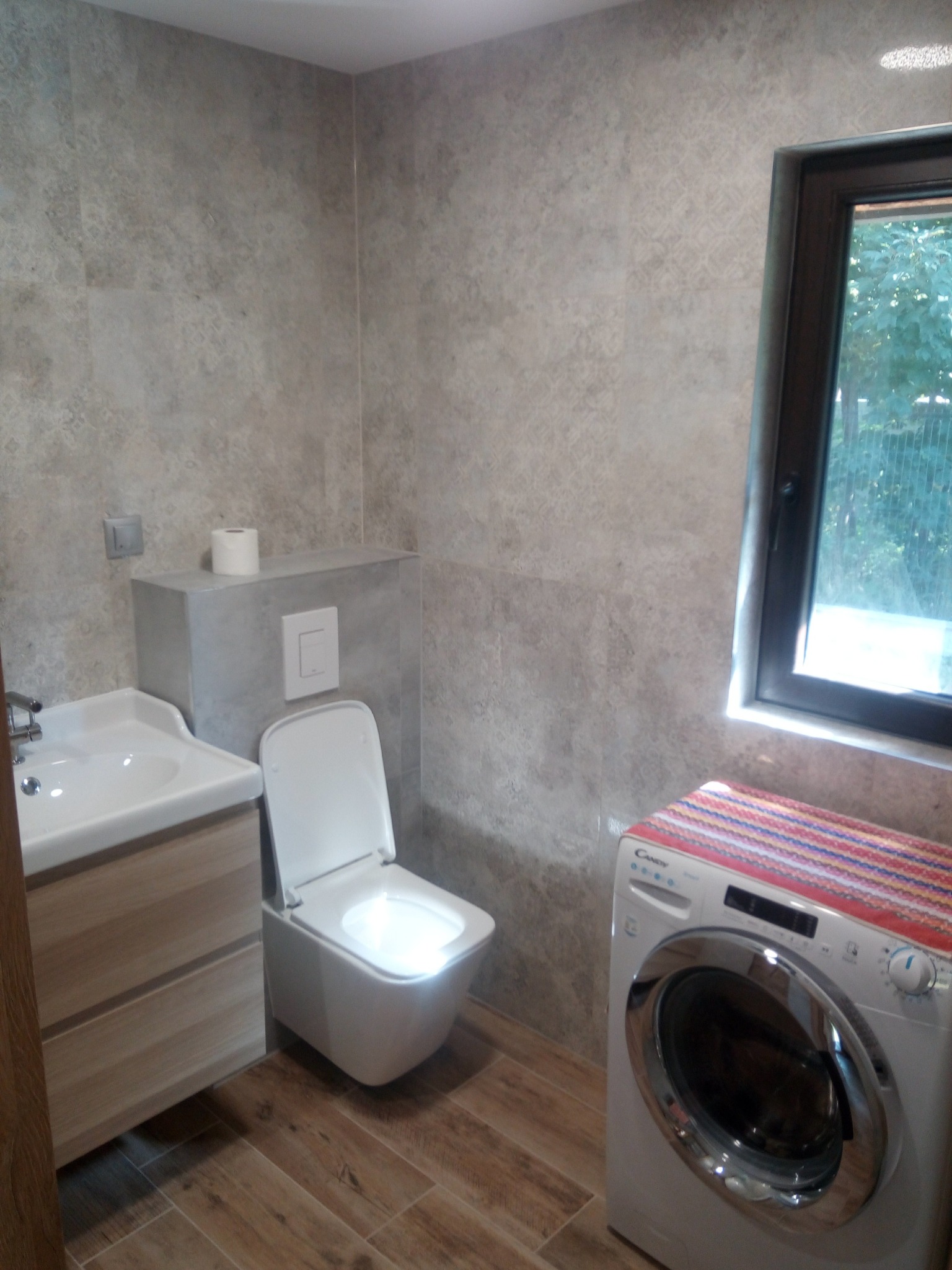 Wykończona łazienka z drewnopodobnymi płytkami na podłodze, umywalką z szafką w jasnym drewnie, toaletą i pralką nakrytą kolorową narzutą przy oknie.