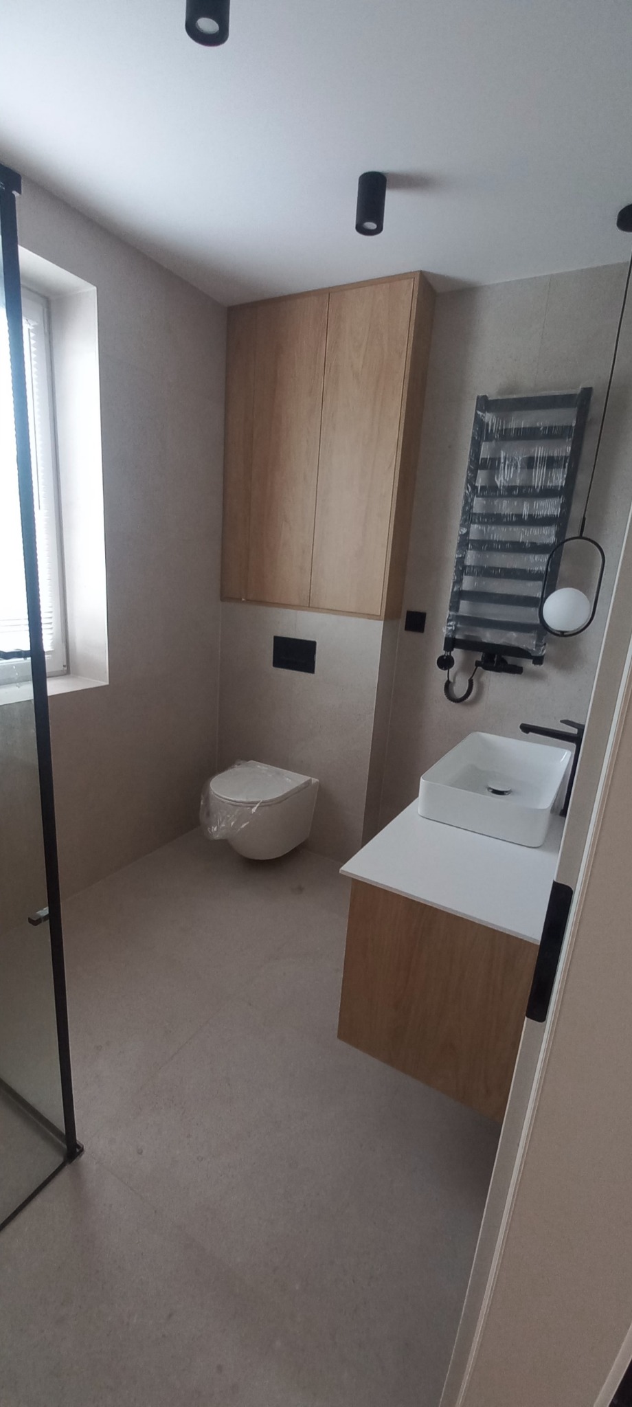 Nowoczesna łazienka po remoncie: beżowe płytki, podwieszana toaleta z folią ochronną, drewniana szafka nad nią, grzejnik drabinkowy z folią, biała umywalka.