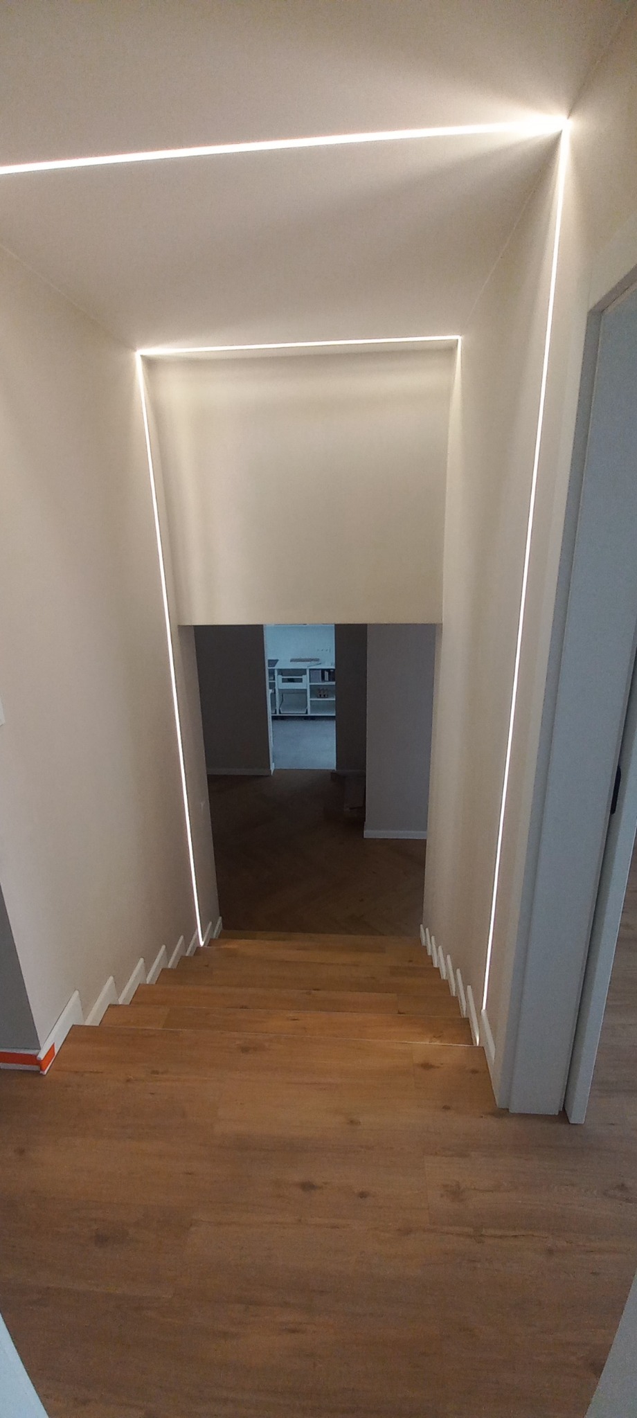 Schody z drewnianymi panelami i nowoczesnym oświetleniem LED wzdłuż ścian, prowadzące do jasnego pomieszczenia. Minimalistyczny design.