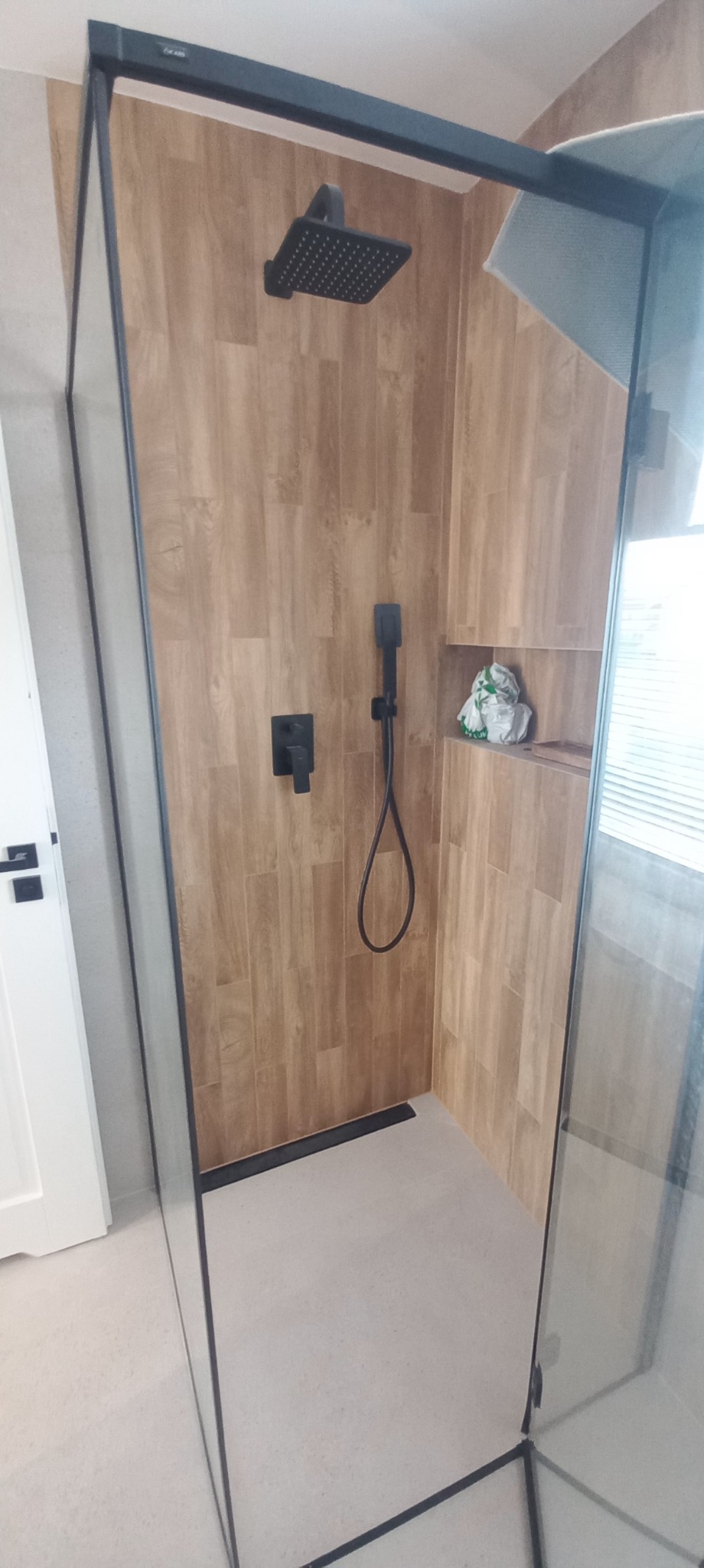 Nowoczesna kabina prysznicowa z deszczownicą i panelami imitującymi drewno. Czarna armatura, minimalistyczny design, jasna podłoga. Wykończenie łazienki w neutralnych barwach.