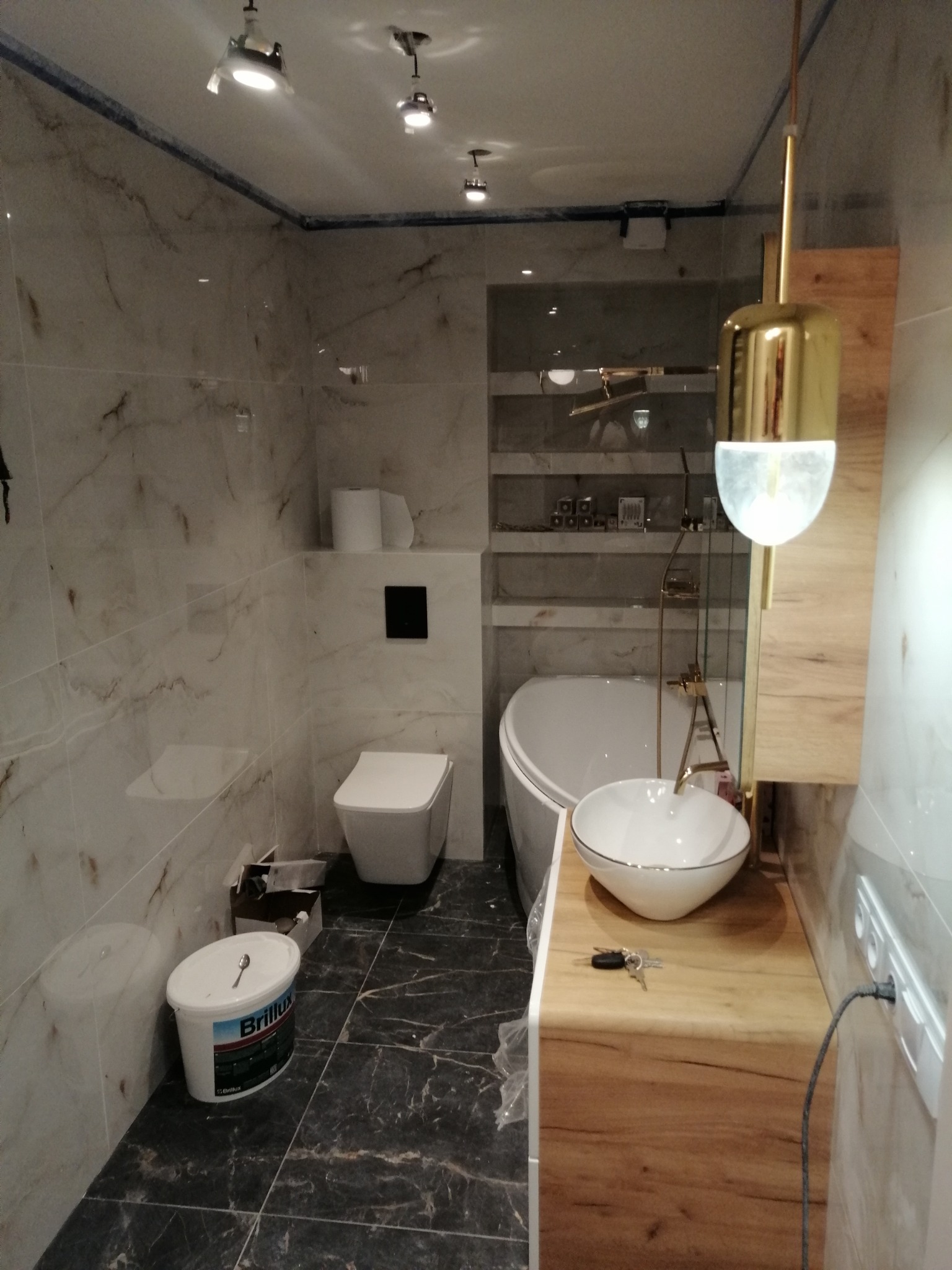 Wykończona łazienka w marmurowej stylistyce z wanną, umywalką na drewnianym blacie i wiszącą toaletą. Widoczny pojemnik z farbą i designerska lampa.