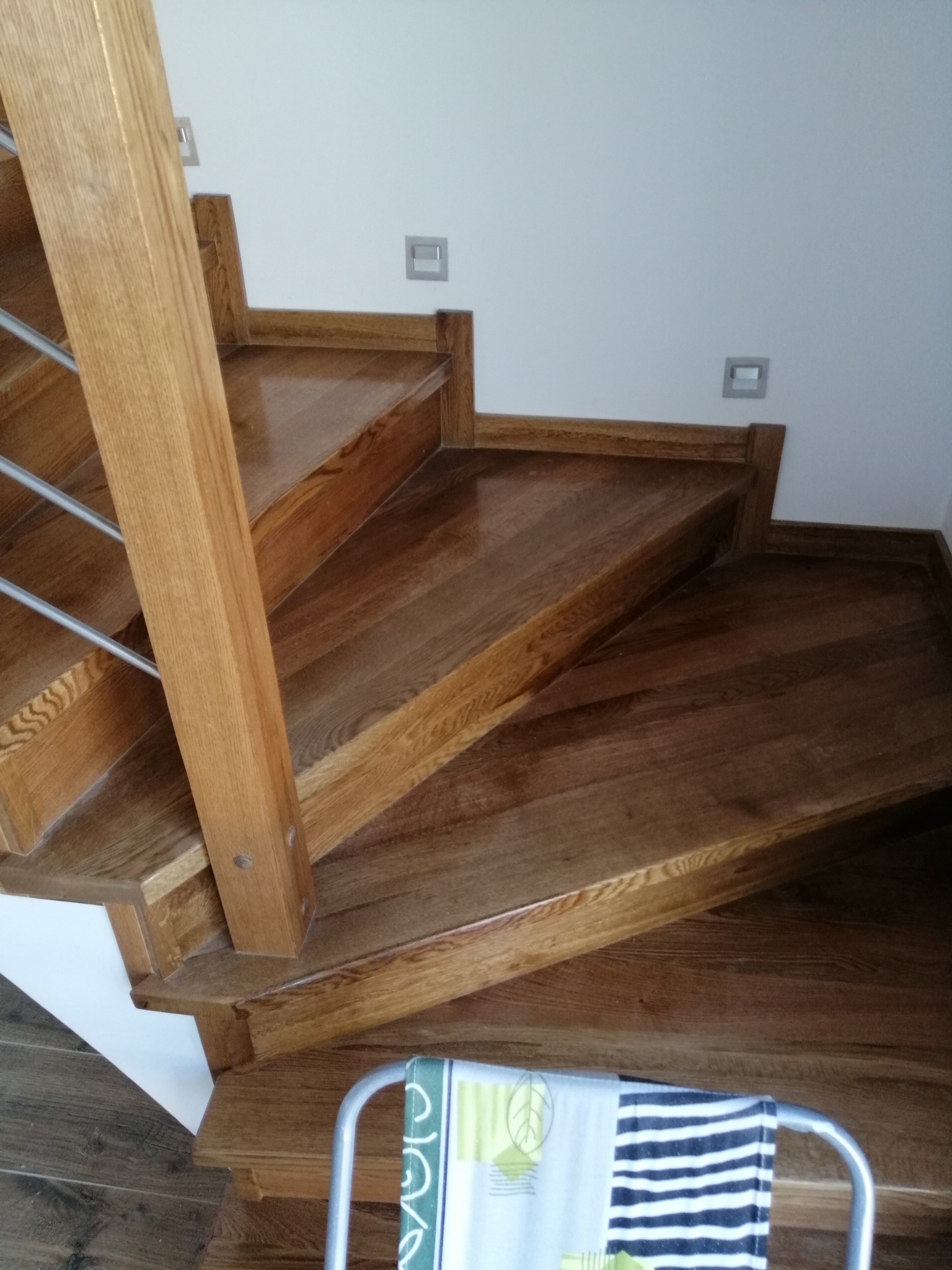 Drewniane schody dębowe z metalową balustradą, oświetlone punktowo, widok z góry. Na dole fragment krzesła ze wzorzystą tkaniną.