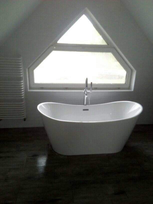 Nowoczesna, wolnostojąca wanna na tle ciemnej podłogi i trójkątnego okna na poddaszu. Minimalistyczny design, grzejnik drabinkowy z lewej strony.