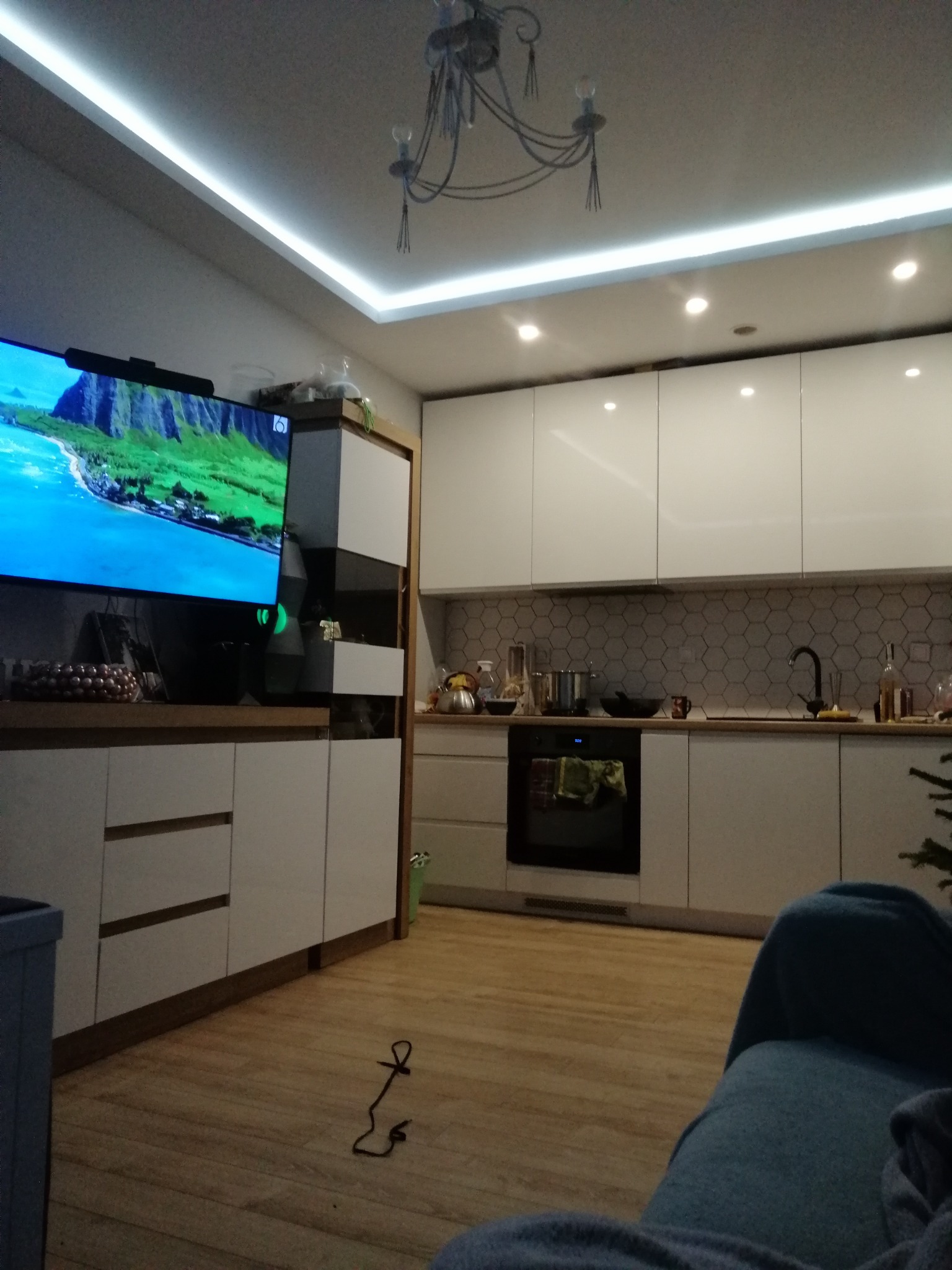 Nowoczesna kuchnia z białymi szafkami, heksagonalnymi płytkami na ścianie i oświetleniem LED. Telewizor z widokiem na ocean. Parkiet na podłodze.