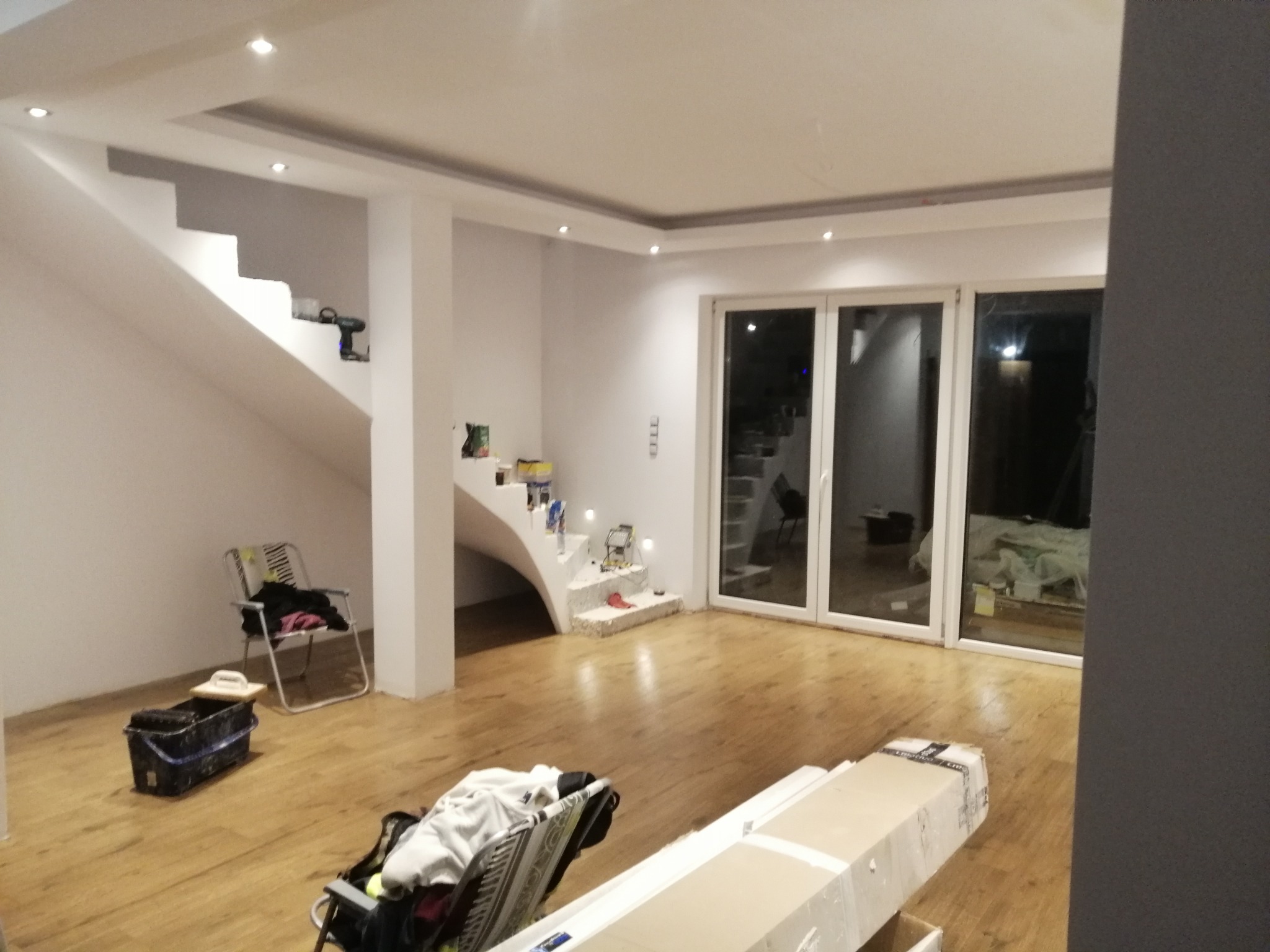 Jasne wnętrze po remoncie z drewnianą podłogą, schodami z oświetleniem punktowym i dużymi oknami balkonowymi. Widoczne narzędzia malarskie.