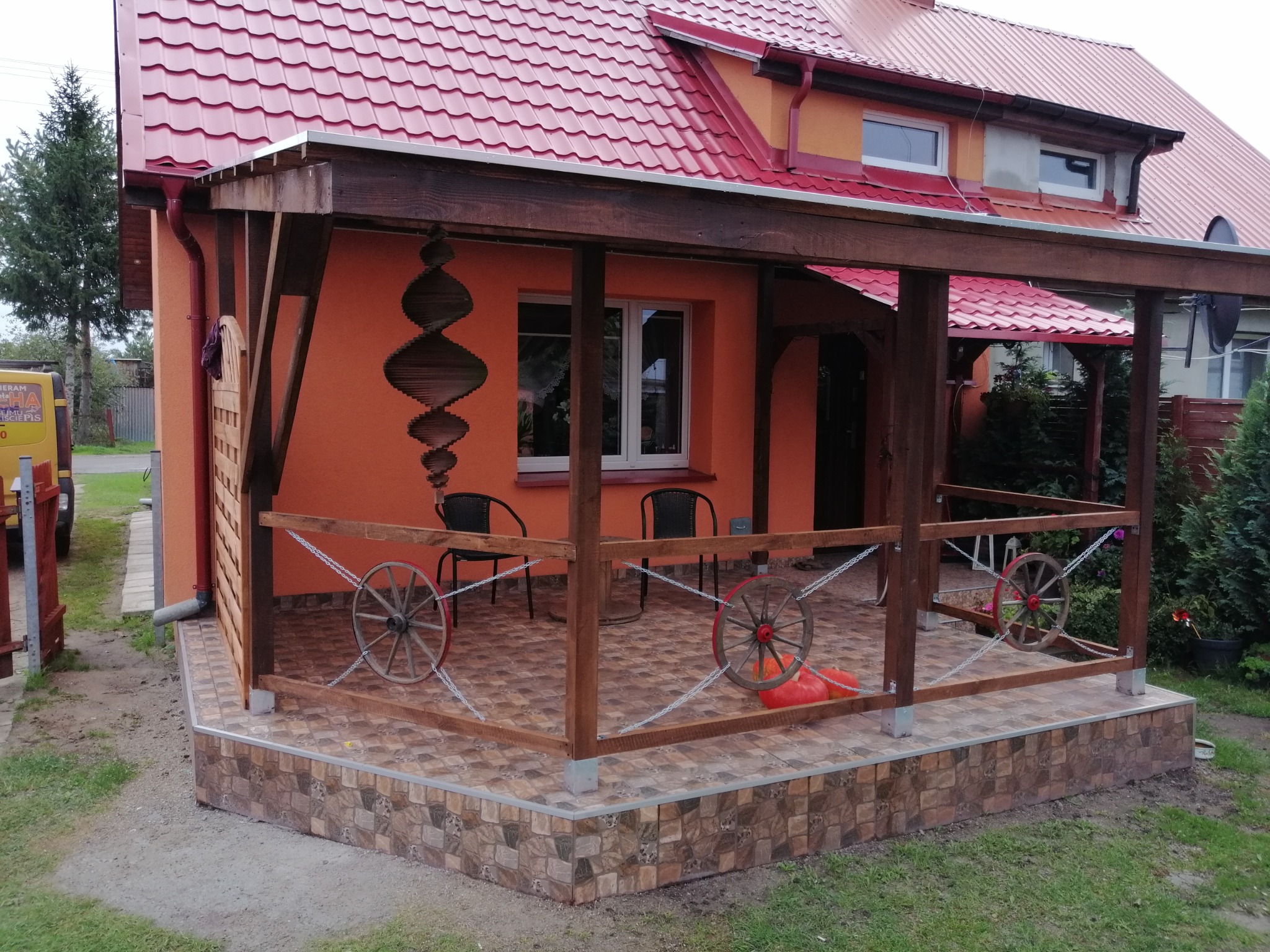 Pomarańczowy dom z zadaszonym tarasem wyłożonym płytkami imitującymi kamień. Dekoracyjne koła od wozu przy barierce i spiralna ozdoba na ścianie.