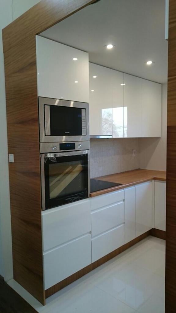 Nowoczesna kuchnia z białymi, lakierowanymi frontami szafek i drewnianymi elementami wykończeniowymi, oświetlona punktowymi lampami sufitowymi, z zabudowanym piekarnikiem i mikrofalówką.
