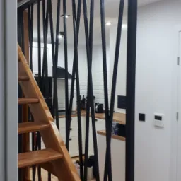 Schody drewniane z balustradą metalową o geometrycznym wzorze, widok na jasną kuchnię z białymi meblami i drewnianym blatem.