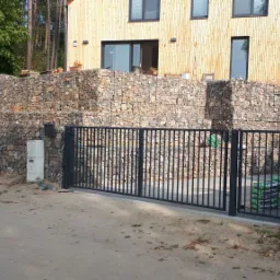 Nowoczesna, metalowa brama wjazdowa na posesję z kamiennym murem gabionowym w tle, widoczny pracownik podczas montażu, narzędzia i worki z cementem.