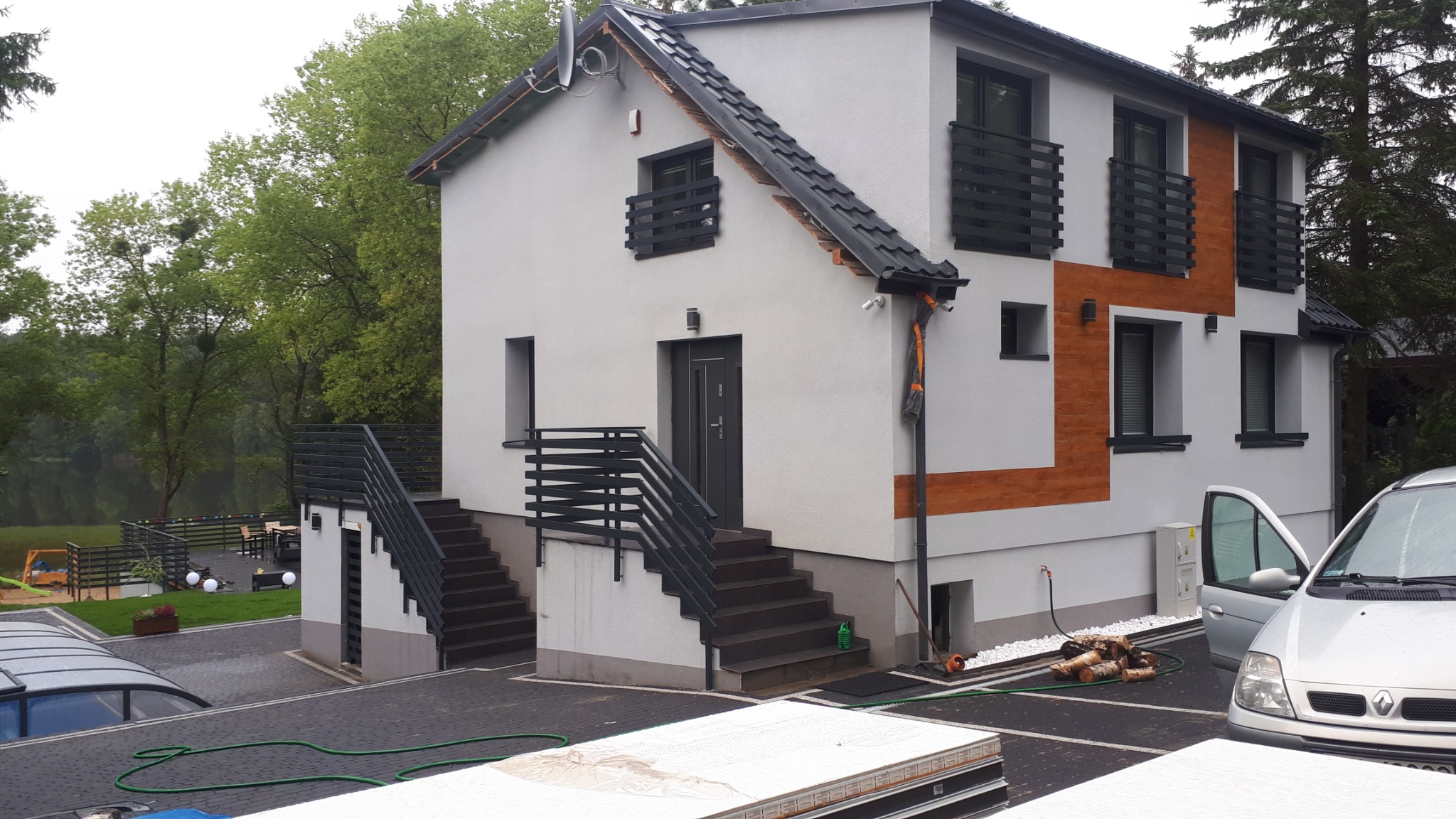 Nowoczesny dom z szarą elewacją, czarnymi balustradami balkonowymi i schodami zewnętrznymi z poręczami, położony w Bieszkowicach, z widokiem na jezioro w tle.