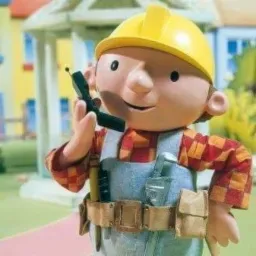 Figurka Boba Budowniczego w kombinezonie, kasku i koszuli w kratę, trzymającego krótkofalówkę, z narzędziami przy pasie, na tle rozmytego ogrodu i domu.