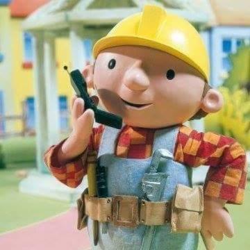 Figurka Boba Budowniczego w kombinezonie, kasku i koszuli w kratę, trzymającego krótkofalówkę, z narzędziami przy pasie, na tle rozmytego ogrodu i domu.