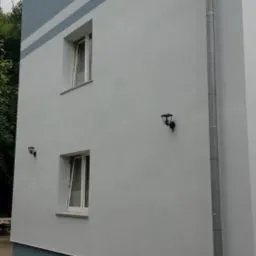 Dwukondygnacyjny budynek z nową, jasnoszarą elewacją i niebieskimi akcentami, widoczna rynna spustowa, okna z białymi ramami i fragment chodnika.