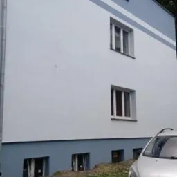 Odnowiona elewacja budynku wielorodzinnego w odcieniach bieli i błękitu, z widocznym fragmentem zaparkowanego srebrnego samochodu osobowego na pierwszym planie.