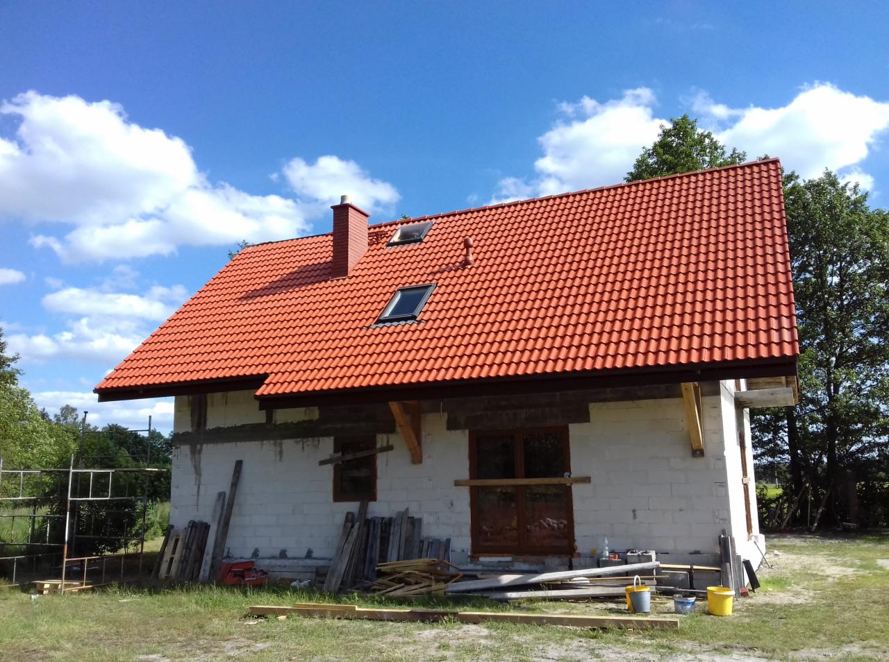 Czerwony dach z dachówką ceramiczną na niedokończonym domu z widocznymi oknami dachowymi i kominem, otoczony zielenią i błękitnym niebem z chmurami.
