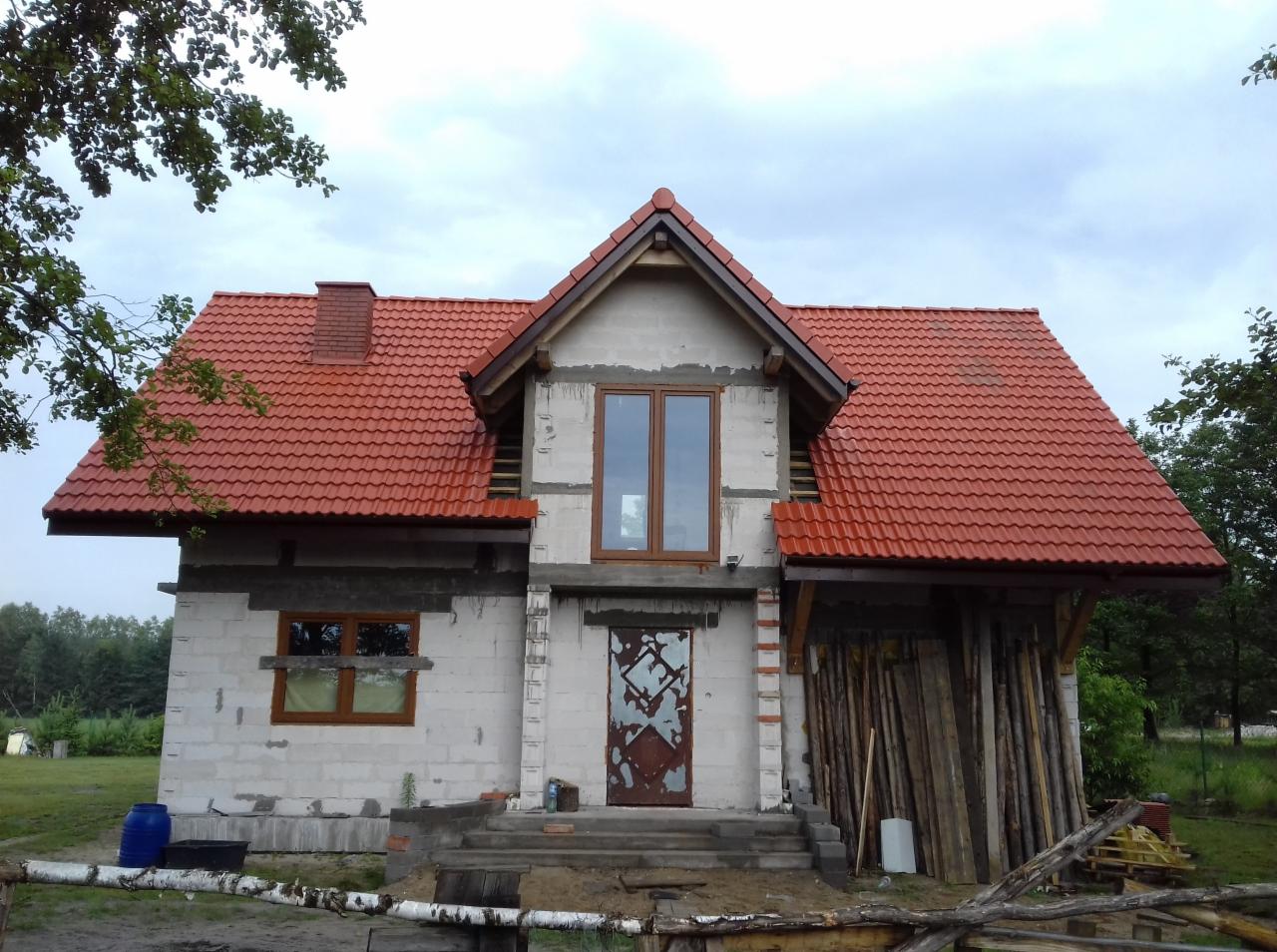 Dom w trakcie budowy z nowym, czerwonym dachem z dachówki ceramicznej, widoczne niedokończone obróbki blacharskie i drewniane konstrukcje wsporcze przy wejściu.