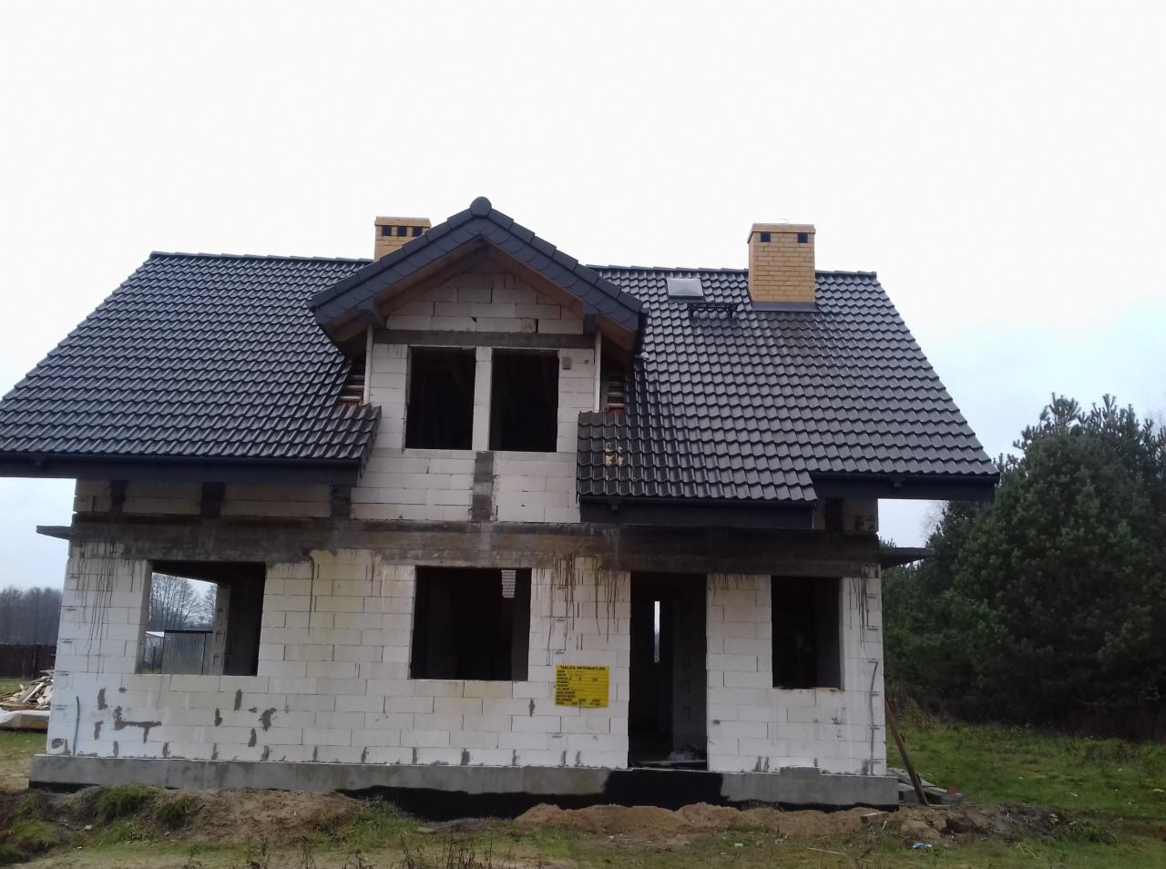 Dom w trakcie budowy z czarnym dachem z dachówki ceramicznej, widoczne kominy z cegły klinkierowej i okno dachowe, ściany z białego bloczka.
