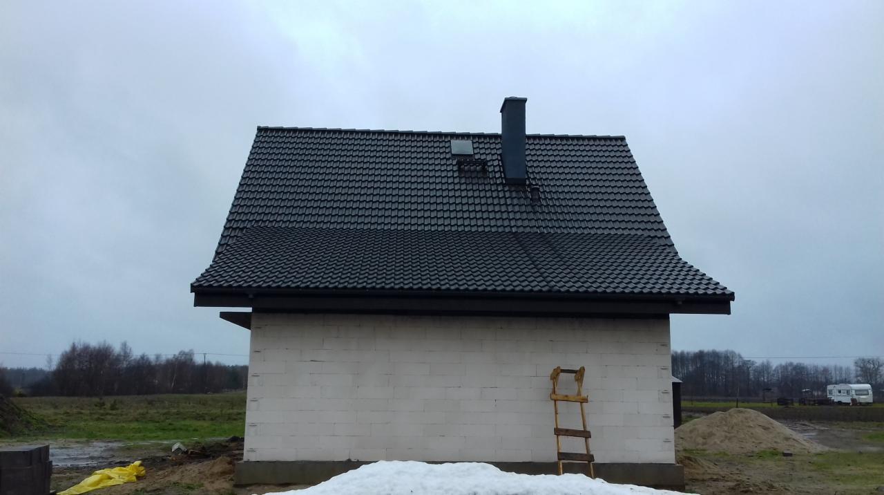 Surowy stan domu jednorodzinnego z ciemną dachówką, widoczny komin, okno dachowe, oraz drewniana drabina oparta o ścianę. Na pierwszym planie resztki śniegu, w tle zalesiony teren.