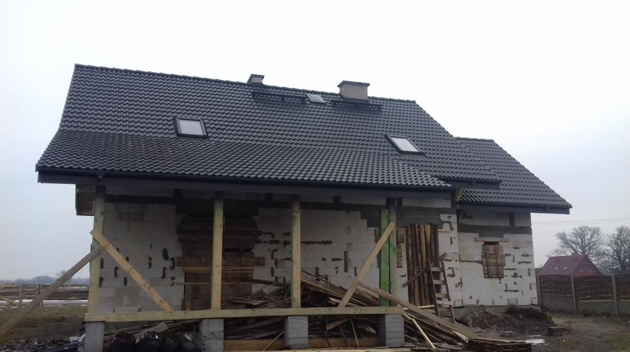 Dom w trakcie budowy z ciemną dachówką ceramiczną, widoczne okna dachowe i kominy, ściany z pustaków, drewniane podpory i rusztowania.