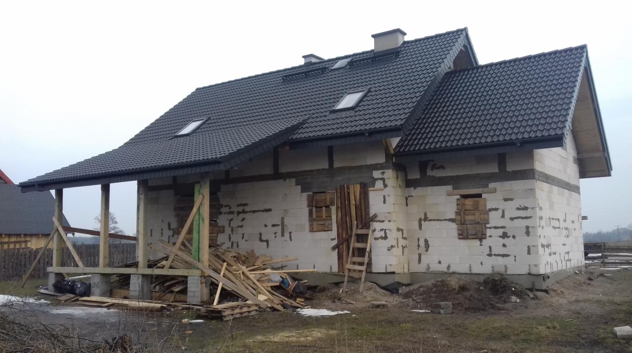 Niedokończony dom z ciemnoszarą dachówką, widoczne okna dachowe i komin, ściany z białych bloczków, drewniane deski zabezpieczające okna, sterta drewna na werandzie, pochmurne niebo w tle.