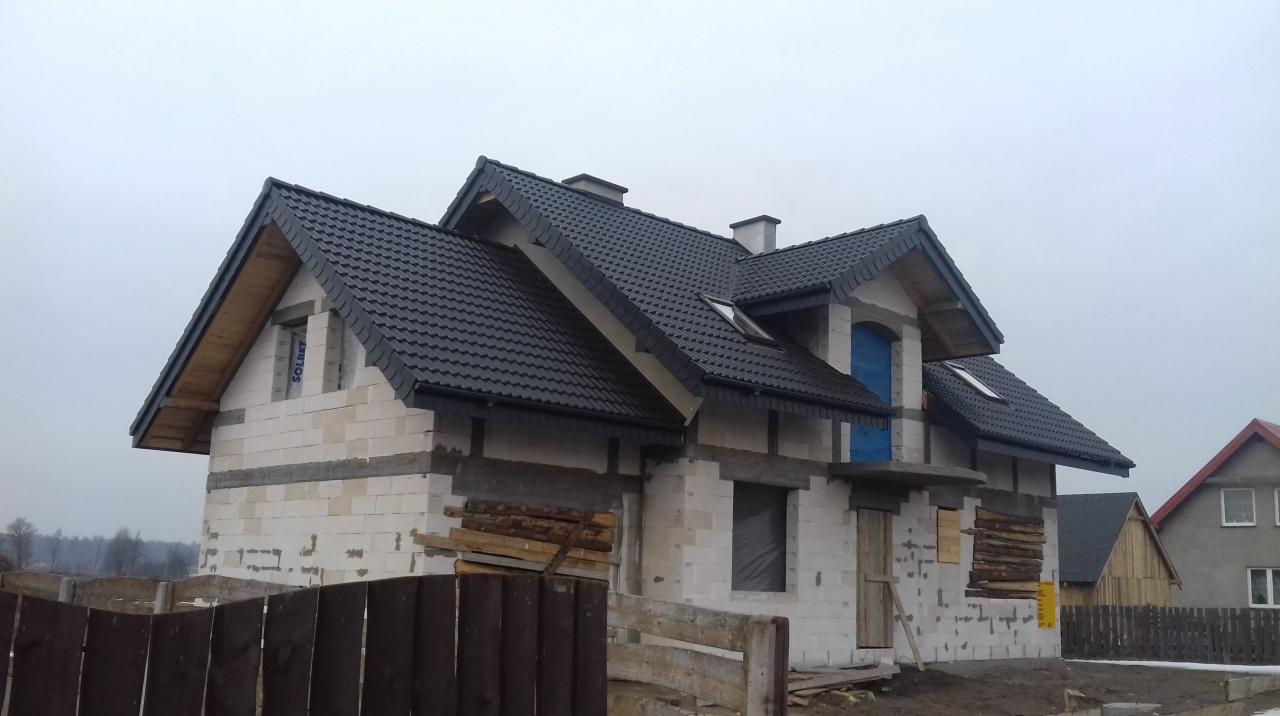 Dom w trakcie budowy z ciemnoszarą dachówką, widoczne kominy i okna dachowe, ściany z białych bloczków, drewniane deski zabezpieczające otwory okienne, pochmurne niebo w tle.
