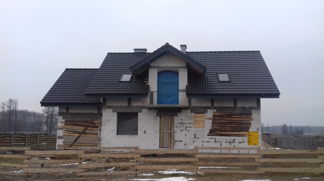 Dom w trakcie budowy z czarną dachówką, zabezpieczonymi oknami i drzwiami, widoczne rusztowanie i tablica informacyjna na elewacji.