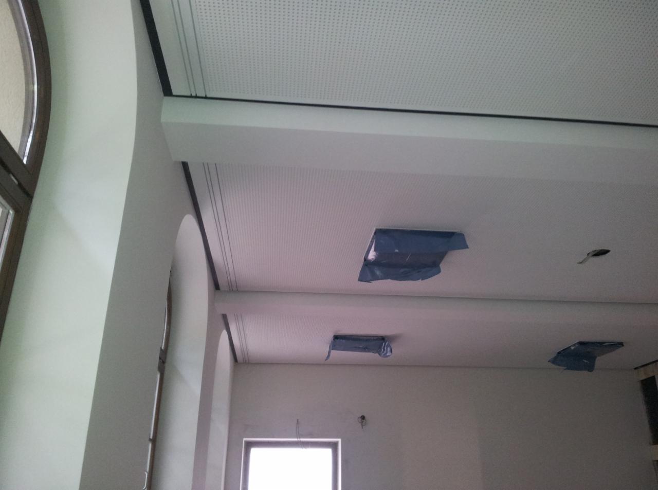 Montaż perforowanego sufitu podwieszanego z widocznymi otworami na oświetlenie zabezpieczonymi folią ochronną, widok z dołu na tle białych ścian z oknem.