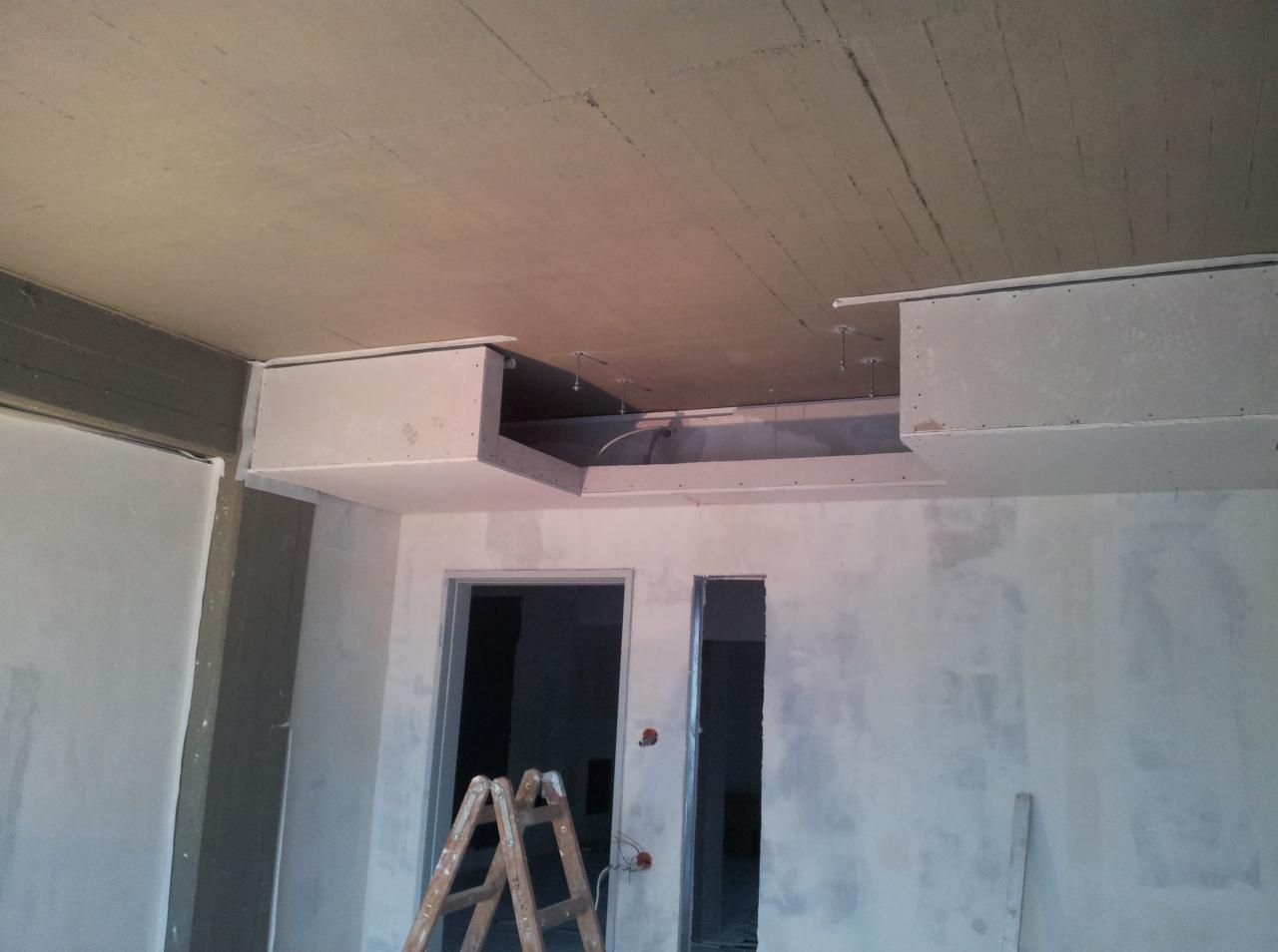 Montaż konstrukcji sufitu podwieszanego z widocznymi profilami i przewodami, surowe ściany po szpachlowaniu, drabina w pomieszczeniu biurowym w trakcie remontu.