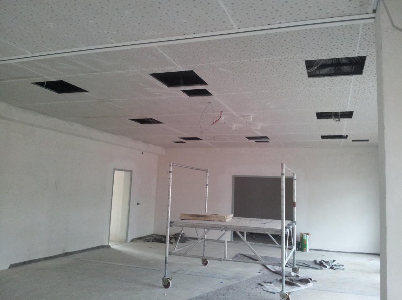 Montaż sufitu podwieszanego z widocznymi otworami na instalacje, rusztowanie na środku pomieszczenia, surowy stan wykończenia ścian i podłogi.